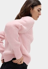 Maglione rosa oversize con finitura textured, polsini e orlo a coste, e silhouette rilassata. Abbinato a jeans scuri.