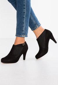 Svarta högklackade ankelstövlar i mjukt mocka, med sidodekoration i elastisk panel och en liten guldbeslag. Bärs med uppvikta denimjeans.