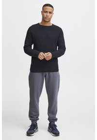 Pull en tricot noir avec col rond, associé à un pantalon de survêtement gris et des chaussures de sport noires. Comprend des poignets et une ceinture côtelés.