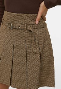 Bruine geruite rok met plooien en een riem, voorzien van een vierkante zilveren gesp. De stof heeft een gestructureerd patroon en een getailleerde taille.