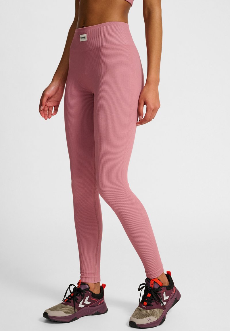 Lichtroze geribde leggings met een hoge tailleband, gecombineerd met donkere sneakers met rode accenten en een grijze zool. Gelaagde afwerking.