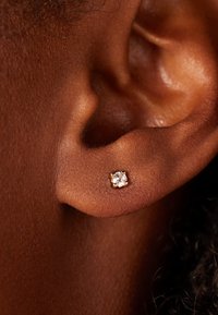 Boucle d'oreille en or avec une gemme ronde et transparente. La boucle d'oreille repose sur un teint de peau foncé, mettant en valeur sa petite taille et son éclat subtil.