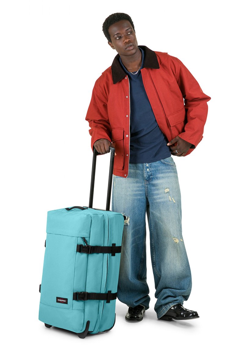 Maleta rodante color verde azulado con correas negras y ruedas; forma rectangular con tela texturizada; acompañada por una persona con chaqueta roja y vaqueros azules.