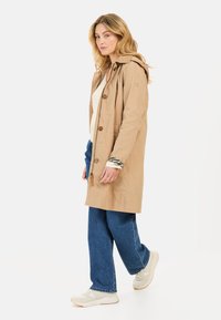 Beige Regenjacke mit Knöpfen und Kapuze, kombiniert mit weiten blauen Jeans und weißen Sneakern. Die Jacke hat eine glatte Textur.
