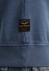 Plava dukserica s rebrastim rubom. Ima crnu flaster s zlatnim natpisom "PME LEGEND" i krilima, s natpisima "AMERICAN CLASSIC" i "TESTED AT HIGH ALTITUDE."