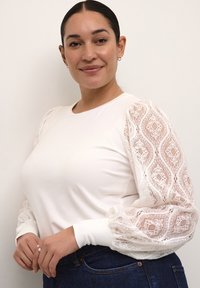 Biely dlhý rukáv s čipkovými balónovými rukávmi s precízne tkanými vzormi, v kombinácii s tmavými džínsami. Hladké bavlnené telo.