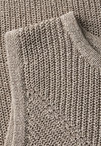 Beige stickad tröja med en strukturerad mönster, rund halsringning, ribbade ärmslut och nederkant, som visar en blandning av öppna och täta stygn.
