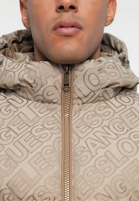 Detailní záběr na osobu oblečenou v béžové mikině na zip s kapucí, vzorovanou opakujícími se slovy "GUESS" a "LOS ANGELES."