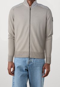 Světle šedý svetr na zip s ribbed texturou a kontrastními ramenními panely. Má vysoký límec a přiléhavé manžety. Spárován s modrými džínami.