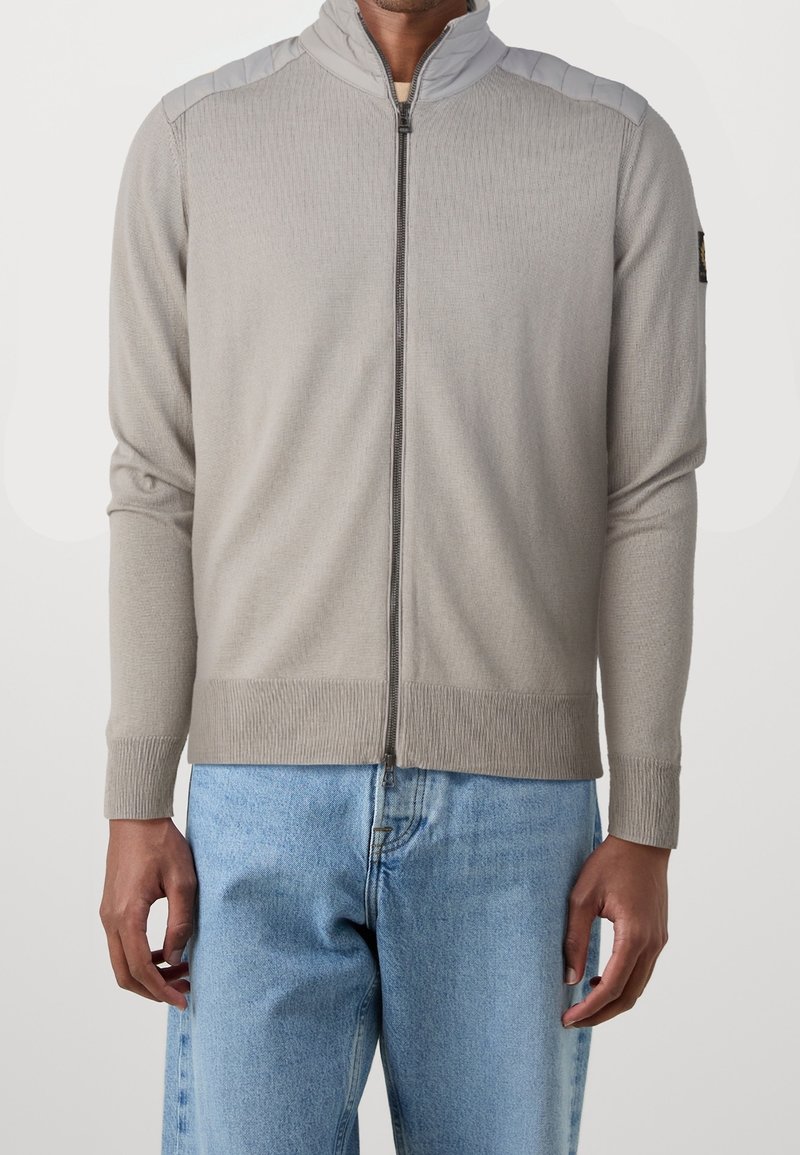 Světle šedý svetr na zip s ribbed texturou a kontrastními ramenními panely. Má vysoký límec a přiléhavé manžety. Spárován s modrými džínami.