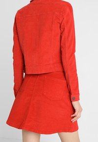 Giacca di velluto a coste rossa con design corto e gonna coordinata. Entrambi presentano una superficie testurizzata e accenti minimalisti, comprese due tasche posteriori.