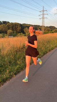 Ženska v črnih tekaških oblačilih teka po asfaltirani poti ob visokem travniku in drevesih med sončnim zahodom pod električnimi napeljavami.