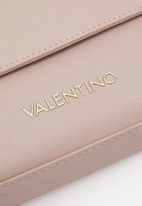 Gaiši rozā teksturēta maku ar zelta "VALENTINO" logotipu. Tam ir gluda apdare un diskrēta izšūšana gar malām.