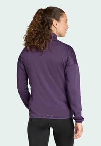 Giacca sportiva viola con maniche lunghe, colletto alto e tessuto testurizzato. Presenta un design aderente e un dettaglio con logo vicino all'orlo.