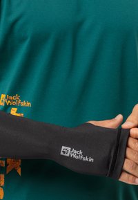 Schwarze Armsocke aus elastischem Material, körpernahe Passform, mit einem Logo am Bündchen. Getragen über einem strukturierten, türkisfarbenen Shirt.
