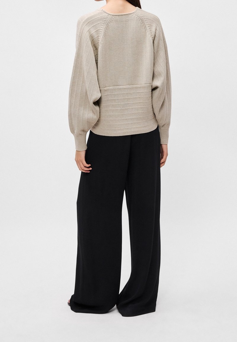 Femme vue de dos portant un pull côtelé beige avec des manches ballon et un pantalon noir à jambes larges sur fond uni.