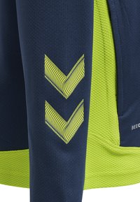Top deportivo azul marino con tejido texturizado, diseño de chevron amarillo brillante en la manga y una inserción de malla en el costado para mayor transpirabilidad.