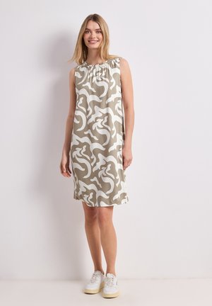 Junge Frau steht an einer weißen Wand, trägt ein ärmelloses beige-weißes Kleid mit abstraktem Muster und weiße Sneakers, lächelnd.