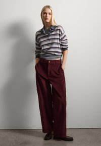 Maglione a righe con tonalità grigie e borgogna, colletto con bottoni; pantaloni in corduroy borgogna larghi con tasche frontali; indossato con scarpe scure.