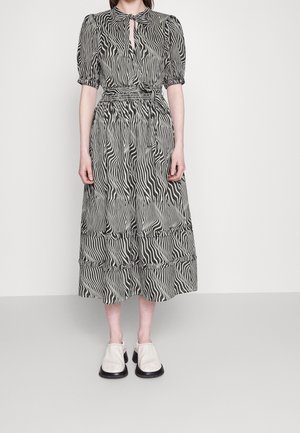 Frau trägt ein schwarz-weißes Midi-Kleid mit Zebramuster, Puffärmeln und weißen Slip-On-Schuhen und steht vor einem einfarbigen Hintergrund.