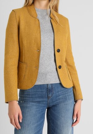 Blazer en laine jaune moutarde avec un col rond, deux poches avant et trois boutons noirs, assorti à un pull gris et un jean bleu.