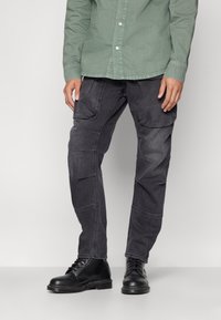 Svarta, strukturerade cargobyxor med flera fickor och en smal passform, parat med svarta snörstövlar och en grön skjorta med knappar.