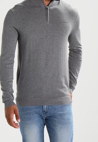 Homme portant un pull gris à manches longues avec une fermeture éclair sur le quart supérieur et des poignets côtelés, associé à un jean bleu clair, sur un fond uni.