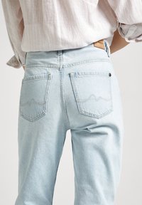 Persona que lleva pantalones de mezclilla azul claros y una camisa de color claro de manga larga con las mangas arremangadas, vista desde la cintura hacia abajo.