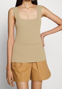 Beige ribbad ärmlös topp med en fyrkantig ringning, åtsittande liv och en fåll som sträcker sig något förbi midjan, i kombination med khaki-shorts.