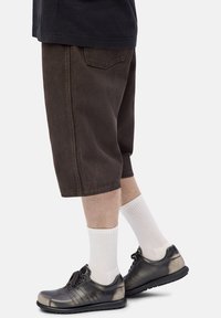 Piernas con pantalones cortos marrón oscuro hasta la rodilla, calcetines blancos de canalé y zapatos negros de cuero con cordones y un ligero brillo.