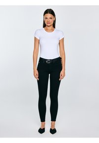 Kobieta stojąca prosto, ubrana w białą koszulkę z krótkim rękawem, czarne obcisłe spodnie z paskiem oraz czarne, spiczaste baleriny na jednolitym tle.