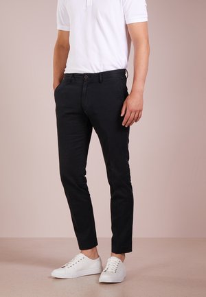 Homme portant un pantalon noir ajusté, un polo blanc et des baskets blanches, debout avec les mains détendues le long du corps sur un fond neutre.