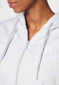 Sweatshirt de capuz cinza claro com um acabamento texturizado, apresentando um capuz com cordão e um logótipo discreto no peito. Fecho e tecido na mesma cor.
