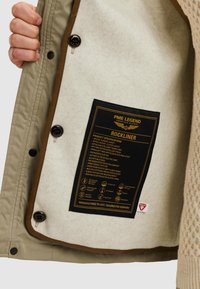 Beige och brun huvad jacka med ett ylleliknande foder. Inuti etiketten finns produktens specifikationer i svart och gul text, med vårdsymboler.