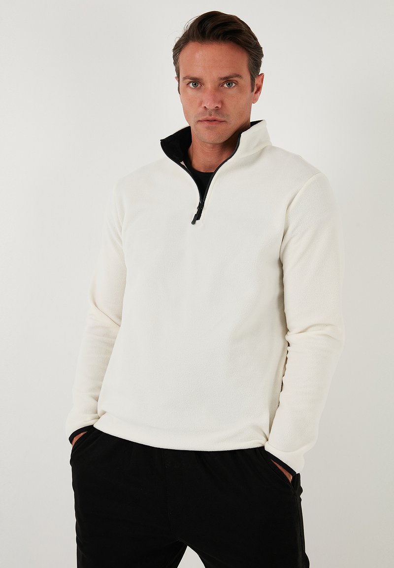 Hvid fleece pullover med en sort lynlåskrave, der har lange ærmer og en blød tekstur. Bæres med sorte bukser.