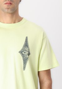 Svetlozelené bavlnené tričko s tmavošedým zdobeným logom na ľavej strane; krátke rukávy, okrúhly výstrih, hladká textúra.