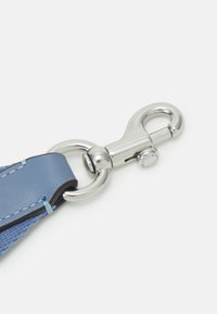 Mousqueton pivotant en métal attaché à un anneau argenté et à une lanière en cuir bleue cousue sur un fond blanc uni.