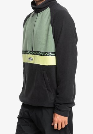 Mann trägt schwarzen und grünen Fleecepullover mit Viertel-Reißverschluss und einem horizontal gemusterten Streifen quer über der Brust.
