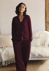 Burgundy knitted cardigan with a V-neck, front buttons, and two pockets, paired with matching pants featuring a relaxed fit and side seams.