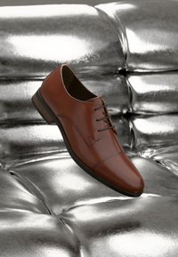 Chaussure habillée en cuir brun avec une finition lisse, design à lacets, bout arrondi et détails de couture subtils, sur fond métallique.