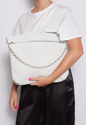 Persona con indossato una maglietta bianca e pantaloni di raso neri che tiene in mano una grande pochette bianca trapuntata con dettaglio a catena dorata.