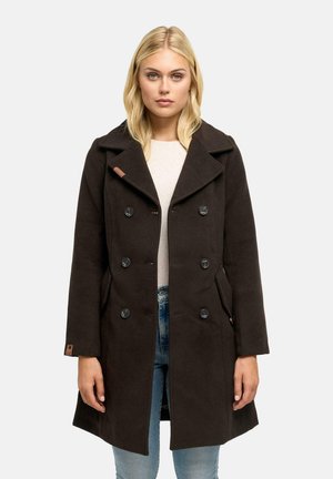Donna che indossa un cappotto nero doppiopetto con ampi revers, abbinato a jeans azzurri chiari e top beige, in piedi davanti a uno sfondo neutro.