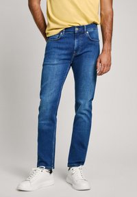 Jean en denim bleu à coupe droite, avec un agencement �à cinq poches, fermeture à bouton et décoloration légère. Associé à des baskets blanches.