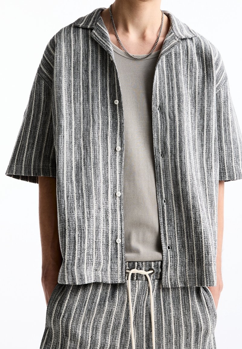 Homme portant une chemise à manches courtes boutonnée à rayures verticales grises et blanches par-dessus un débardeur côtelé beige, accompagné d'un pantalon assorti à rayures avec cordon de serrage.