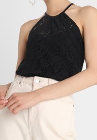 Black lace halter top featuring a textured floral pattern and gathered neckline, paired with high-waisted beige pants.