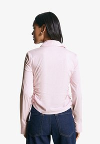 Camicia a strisce rosa chiaro con maniche lunghe, dotata di colletto e dettagli di arricciatura laterale regolabili. Abbinata a jeans in denim scuro.
