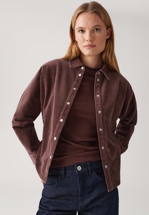 Young woman with straight blonde hair wearing a brown ribbed top under an open brown corduroy button-up shirt and dark denim jeans.