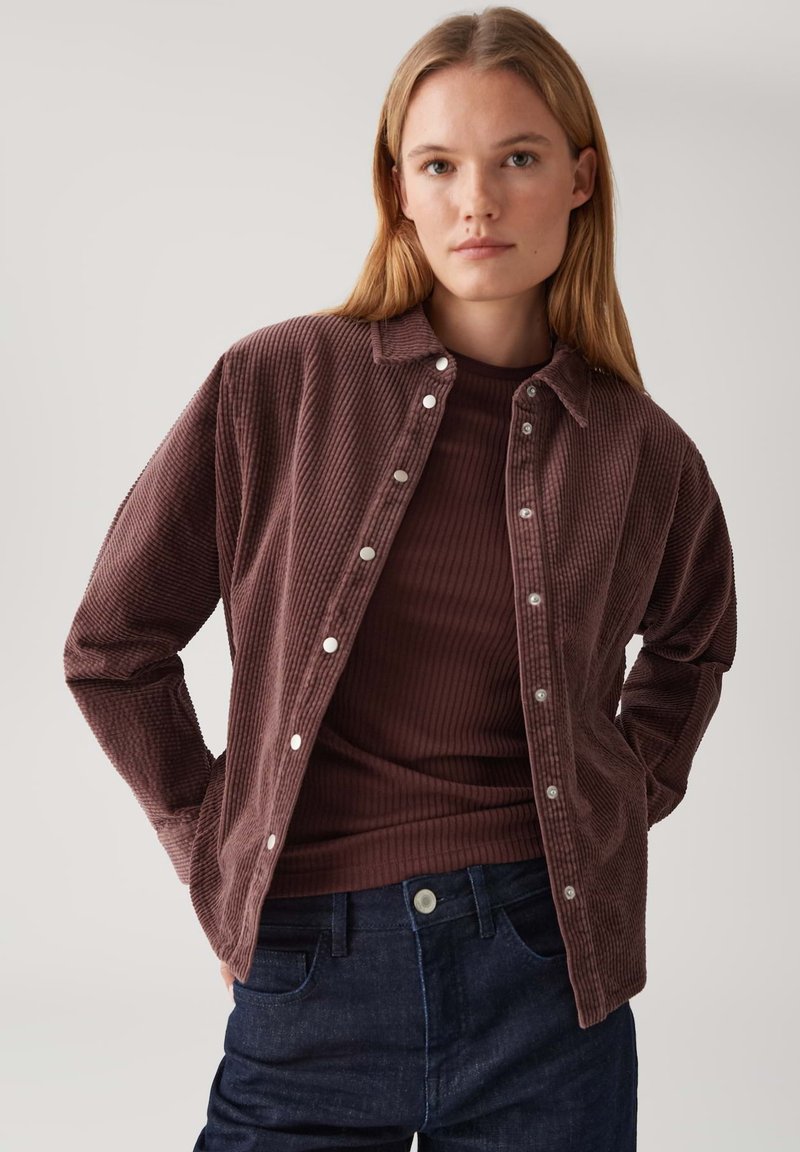 Jonge vrouw met steil blond haar, gekleed in een bruin geribd topje onder een open bruin corduroy overhemd met knopen en een donkere spijkerbroek.