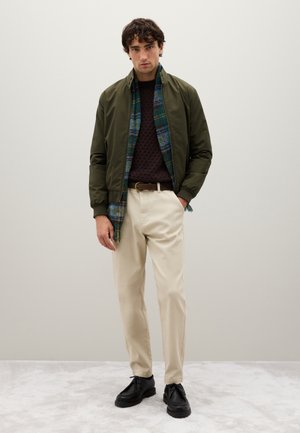 Man in een olijfgroene bomberjack, een donkerbruine trui, beige broek, zwarte schoenen en een blauw-groene geruite sjaal die tegen een effen muur staat.