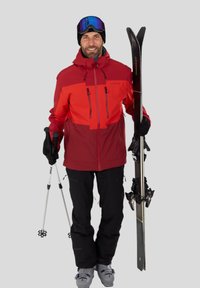 Rote und schwarze Skijacke mit mehreren Taschen, schwarze Skihosen, graue Skischuhe, schwarze Handschuhe, hält Skistöcke und schwarze Skier.